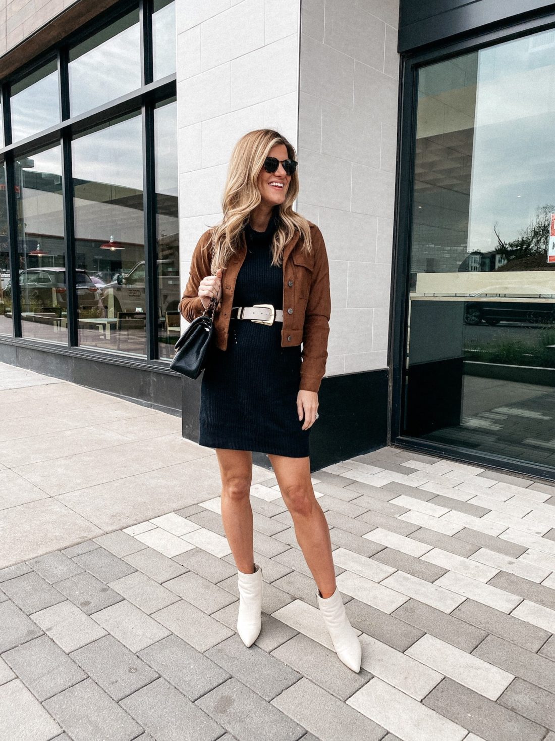 Brighton Butler wearing a black sweater dress, white belt, brown jacket and white booties